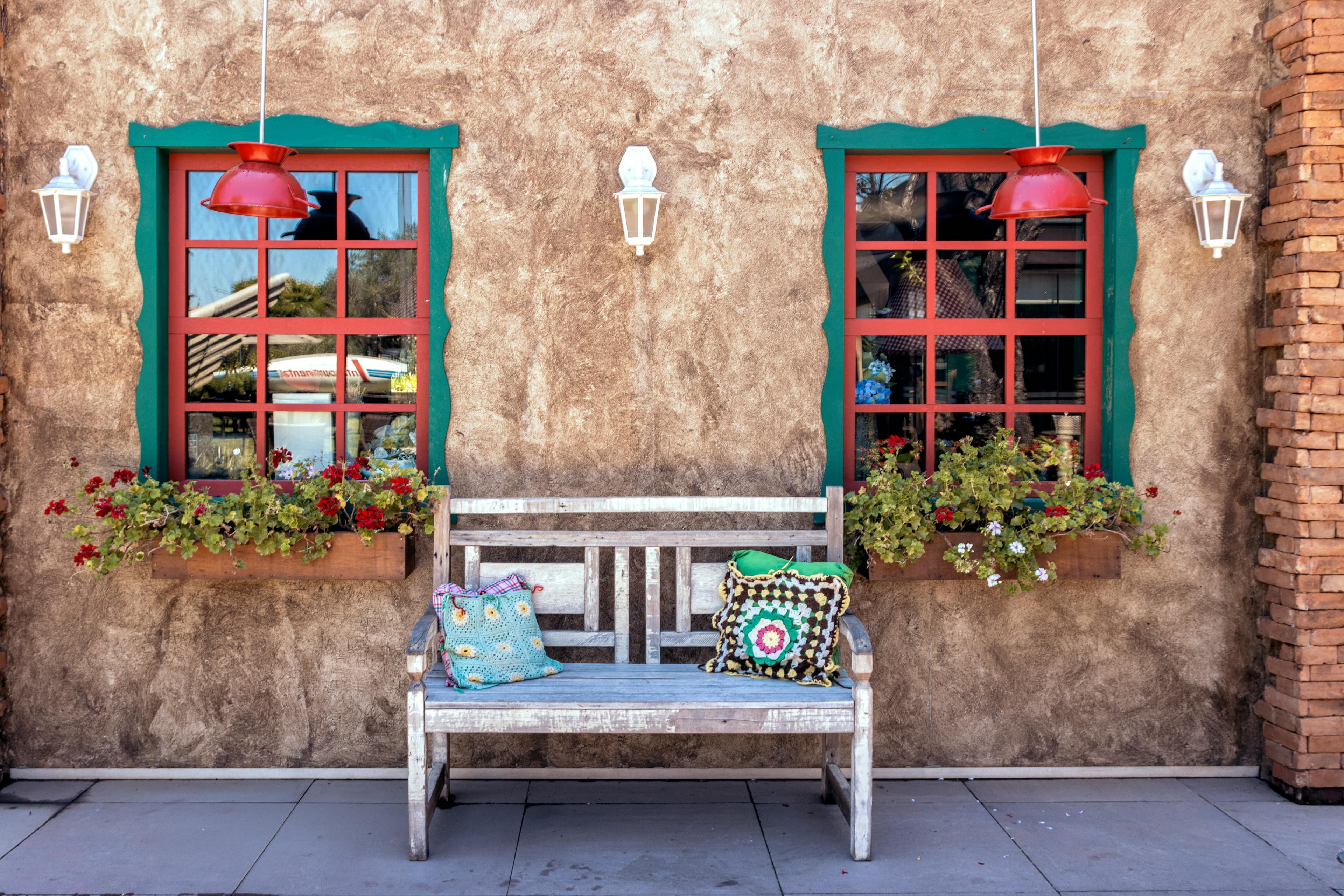 Rustic wooden bench with crochet pillows and window flower boxes, reflecting cozy and colorful summer decor trends.
