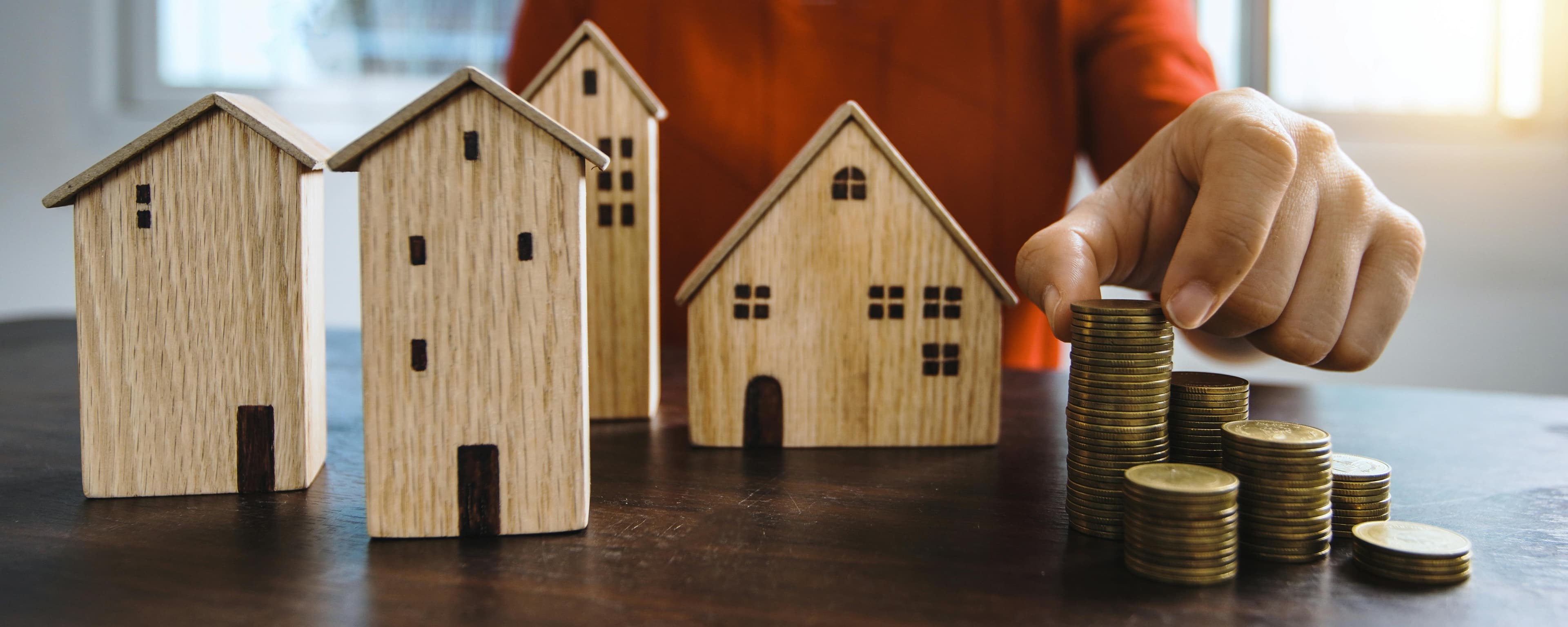 Gemini said A person adds a coin to a growing stack of gold coins, illustrating an investment opportunity next to a row of small wooden house models representing real estate assets.