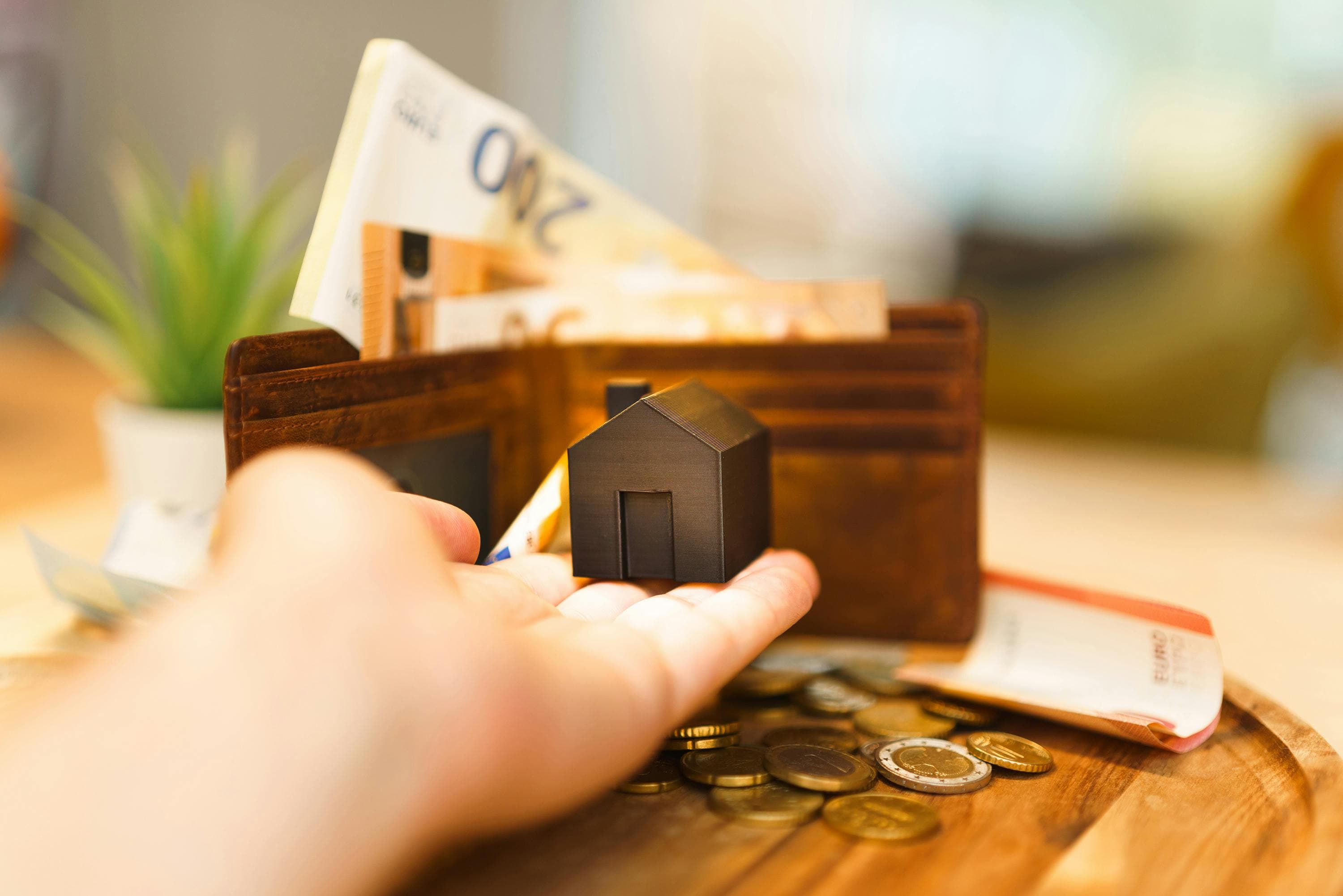 A hand holds a small black model house in the foreground, signifying a prime real estate investment opportunity, with a wallet full of currency notes and scattered coins in the blurred background.