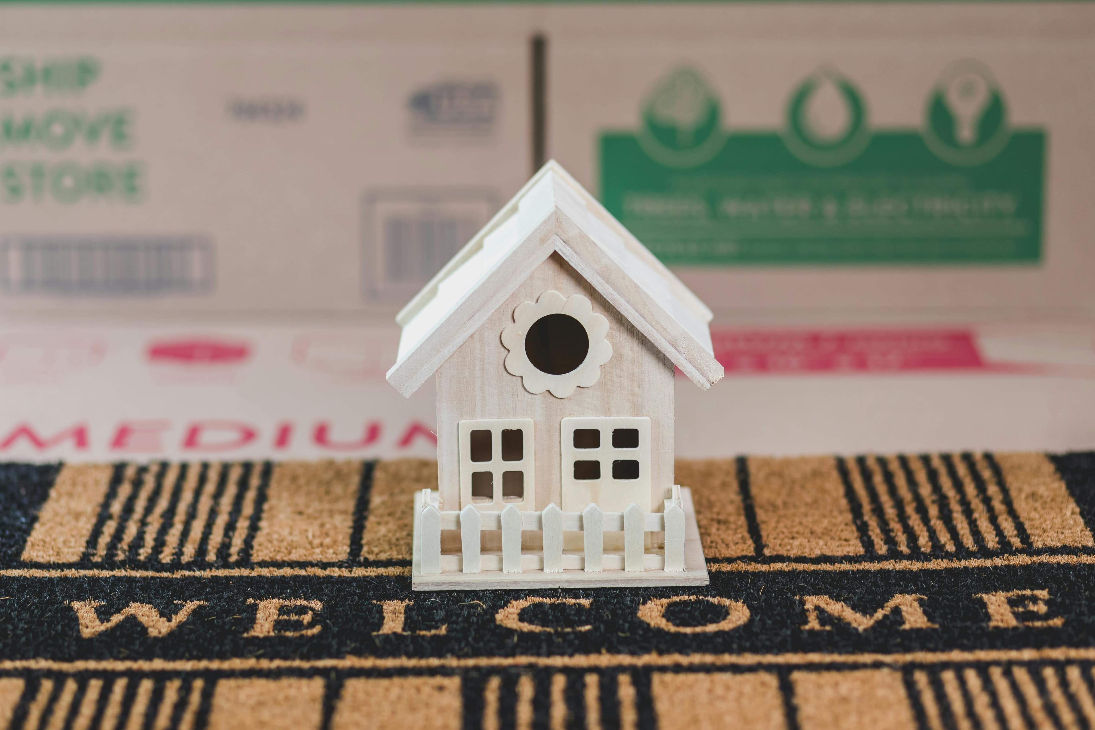 A small, detailed model of a white suburban house sits on a "Welcome" doormat, representing a welcoming investment opportunity in the residential real estate market.